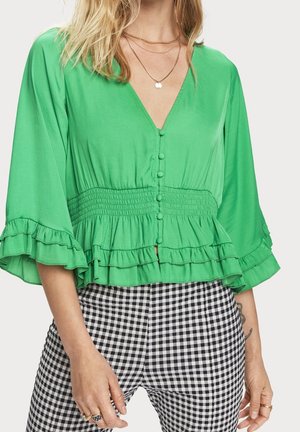 Vrouw die een felgroene blouse met knopen, gerafelde zoom en mouwen draagt, gecombineerd met zwart-witte geruite broek.