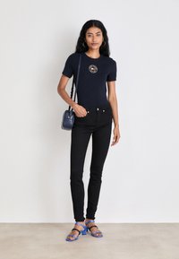 Mørkeblå t-skjorte med kort arm og et rundt logo, svarte skinny jeans, og blå hælsandaler med tynne stropper. Liten crossbody-veske.