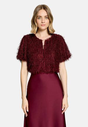 Kvinde med skulderlangt blond hår iført en kortærmet, struktureret bordeaux cardigan over en ærmeløs, tætsiddende bordeaux kjole.