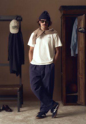 White shirt with logo, brown sweater draped, navy pants, and navy sneakers. Accessories include a navy beanie and sunglasses. Neutral backdrop.