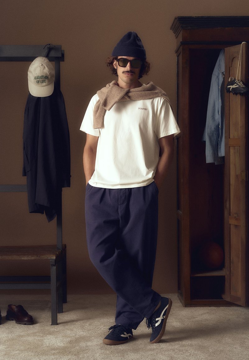 Bijela košulja s logotipom, smeđi pulover preko ramena, tamnoplave hlače i tamnoplave tenisice. Dodatci uključuju tamnoplavi šešir i sunčane naočale. Neutralna pozadina.