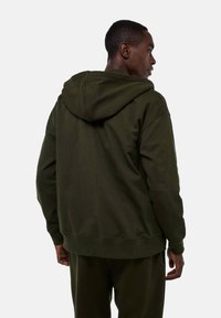Homme de dos, portant un sweat à capuche vert foncé et un pantalon assorti, se tenant contre un fond gris clair uni.