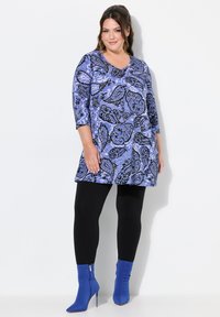 Plus-size tunika s fialovým a černým paisley vzorem, s V-výstřihem a tříčtvrtečními rukávy, spojená s černými legínami a modrými kotníkovými botami.