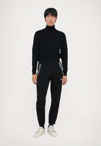 Homem de pé, vestido com um suéter de gola alta preto, calças de treino pretas com zíperes e ténis brancos, em fundo branco liso.