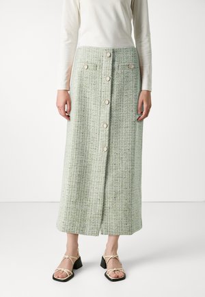 Green tweed midi skirt with textured fabric, front button closure, and side pockets. Paired with beige strappy block-heel sandals.