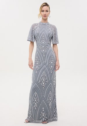 Mujer vestida con un vestido largo plateado-gris con bordado intrincado de cuentas y flores, mangas cortas y un escote alto sobre un fondo liso.