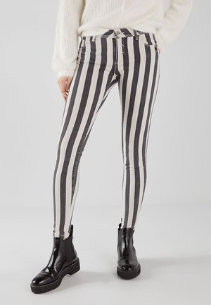 Person iført sort-hvide lodrette stribede skinny jeans, hvid strikket sweater og sorte ankelstøvler, stående på en ensfarvet baggrund.