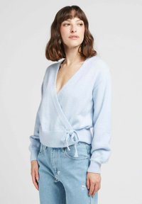 Pull en maille bleu clair à enrouler, fabriqué dans un matériau doux. Il présente un décolleté en V profond, des manches longues et un détail de taille nouée. Associé à un jean clair.
