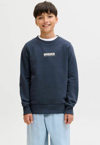 Ragazzo sorridente con capelli corti e scuri che indossa una felpa blu navy con la scritta "BANGKOK" e jeans azzurri, in piedi davanti a uno sfondo bianco semplice.