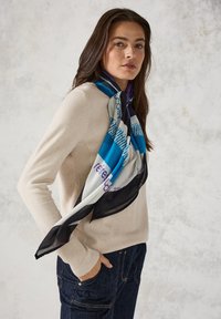 Seidenschal mit geometrischem Design in Blau, Schwarz und Weiß mit lila Akzenten, um den Hals einer Person gewickelt, die einen beigen Pullover trägt.
