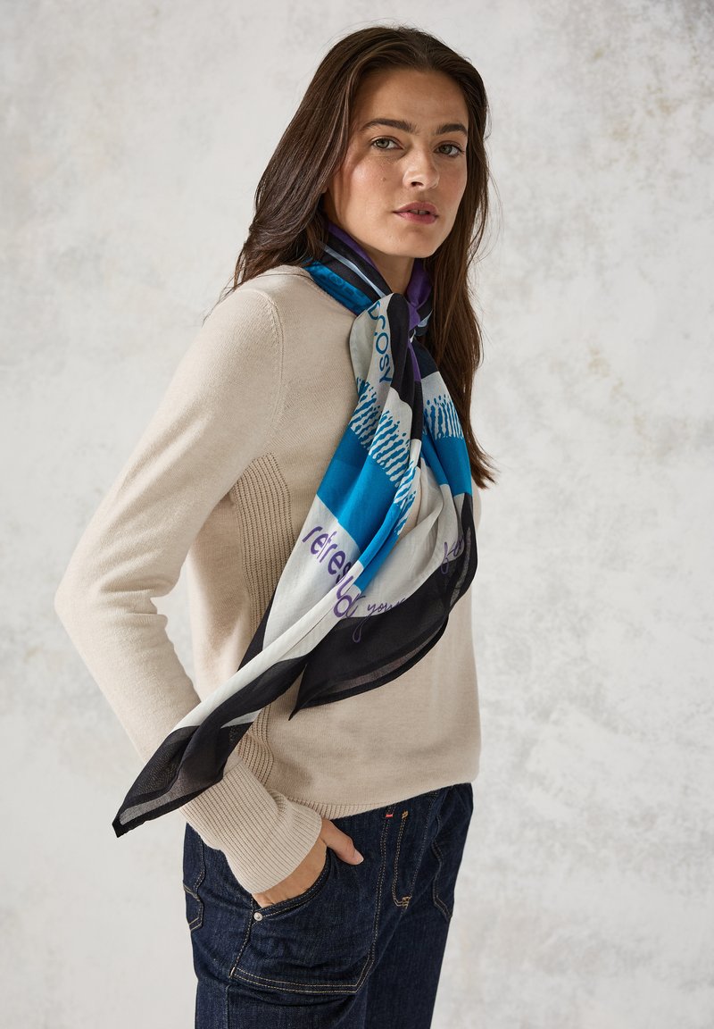 Seidenschal mit geometrischem Design in Blau, Schwarz und Weiß mit lila Akzenten, um den Hals einer Person gewickelt, die einen beigen Pullover trägt.