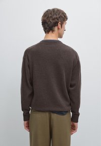 Braun gestrickter Pullover mit Rundhals, langärmlig und mit gerippten Bündchen. Über einem dunkel engen Hemd und olivfarbenen Hosen getragen.