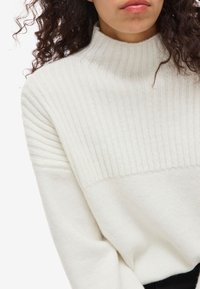 Kremni pleteni pulover z rebrasto teksturo na zgornji polovici in gladko teksturo na spodnji polovici, z ovratnikom.