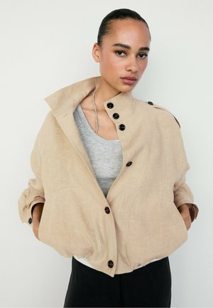 Junge Frau mit zurückgebundenem Haar trägt eine beige, geknöpfte Jacke über einem grauen Shirt, die Hände in den Taschen, vor weißem Hintergrund.