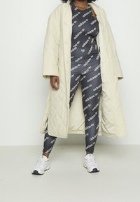 Beżowy pikowany długi płaszcz na dopasowane czarne legginsy z powtarzanym białym nadrukiem logo, w połączeniu z białymi sneakersami z czarnymi akcentami.