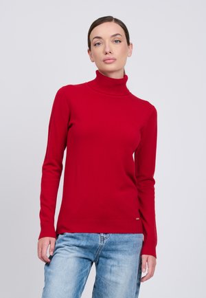 Kvinde iført en tætsiddende rød rullekrave og lyseblå jeans, stående mod en ensartet lys grå baggrund.