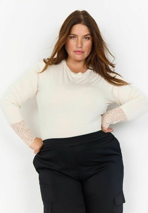Blusa de manga larga color crema con detalles de encaje en los puños, que presenta un escote redondo. Combinada con pantalones negros de cintura alta, de textura suave.