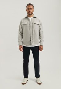Lichtgrijs flanellen shirt met twee voorzakken, zwarte knopen en een kraag, gecombineerd met zwarte broeken en witte sneakers.