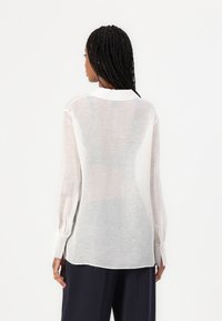 Blusa bianca trasparente con maniche lunghe, colletto classico e polsini con bottone. Il tessuto ha una consistenza leggera e ariosa, mostrando una sottile sfumatura.