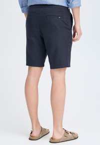 Marineblå shorts laget av lett stoff, med en baklomme med knappelukking. Båret med beige sandaler, som viser nakne bein og føtter.