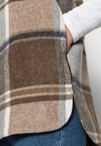 Rutig poncho i brunt och grått med en mjuk textur, featuring en böjd fåll och en sidoficka, bärs över en vit långärmad topp.