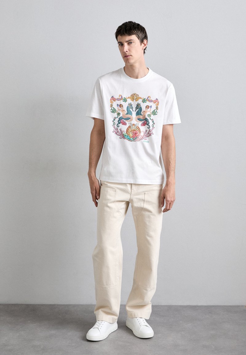 Witte katoenen T-shirt met kleurrijk grafisch ontwerp met figuren en zeemotieven, gecombineerd met lichtbeige broeken en witte sneakers.