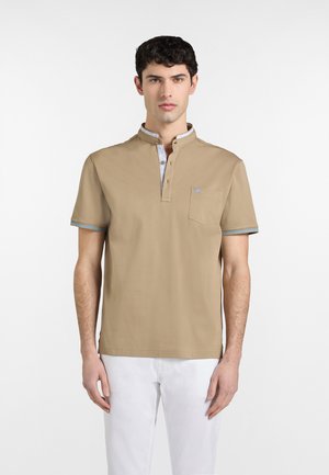 Muž v béžovej polokošeli s krátkymi rukávmi s svetlomodrým lemovaním a bielych nohaviciach, stojaci pred jednoduchým bielym pozadím.