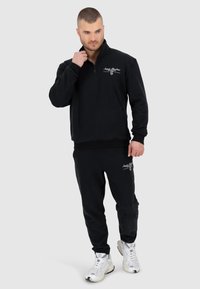 Felpa nera in pile con zip quarti e colletto alto, abbinata a pantaloni della tuta coordinati. Presenta un logo ricamato sul petto e sulla coscia.