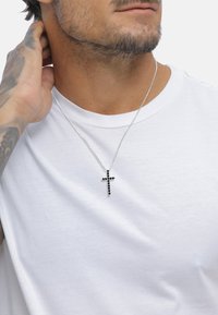 Sølv kædehalssmykke med et sort kors vedhæng pyntet med små sorte sten, båret over en hvid T-shirt.