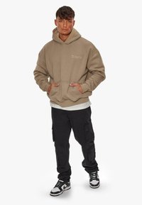 Beige Kapuzenpullover mit einer Kängurutasche und gesticktem Text, kombiniert mit schwarzen Cargo-Hosen und Schwarz-Weiß-Sneakers.