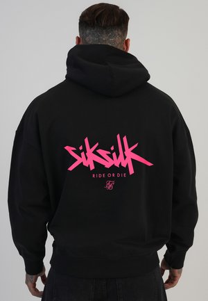 Mężczyzna z tatuażem na szyi ubrany w czarną bluzę z kapturem z jaskraworóżowym napisem „SikSilk Ride or Die” na plecach, zwrócony tyłem do kamery.