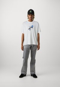 Valge T-särk sinise graafilise disainiga, millele on lisatud hallid teksapüksid tekstuursete detailide ja tõmblukkudega, ning mustad tossud ja müts.