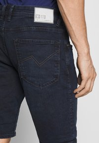 Donker denim shorts met een zichtbare achterzak met een chevron steekpatroon. Metalen knoopaccent in de taille, label op de achterzijde van de tailleband.
