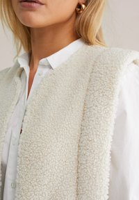 Bela, puhasta, teksturirana brezrokavnik z mehko, puhasto površino, navlečen čez belo srajco s kragnico. Vidna zlatana uhani.