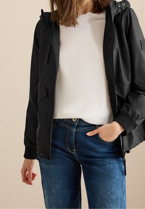 Femme portant une veste noire à capuche sur une chemise blanche et un jean bleu, avec une main dans la poche et un fond neutre.
