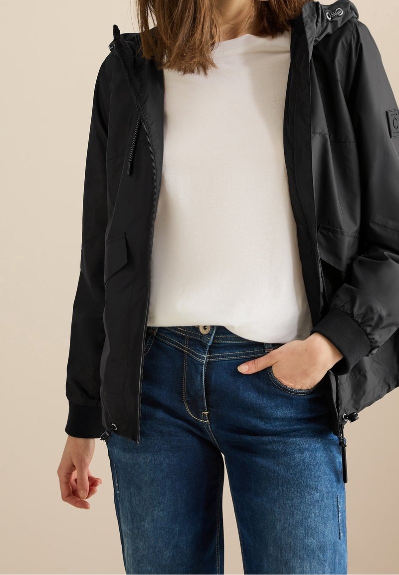 Femme portant une veste noire à capuche sur une chemise blanche et un jean bleu, avec une main dans la poche et un fond neutre.