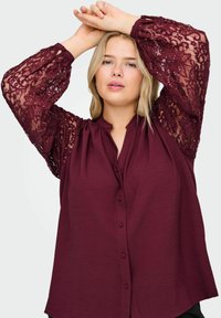 Blusa bordeaux con maniche lunghe in pizzo caratterizzate da motivi floreali e paillettes. Davanti con bottoni e colletto alla coreana. Tessuto stropicciato.