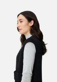 Frau mit langen dunklen Haaren trägt einen hellgrauen Rollkragenpullover unter einem ärmellosen schwarzen, gerippten Pullover und blickt nach links vor einem einfarbigen Hintergrund.