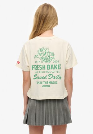Cream cotton crop t-shirt with green graphic design featuring roses and text. Short sleeves and a round neckline, styled with a grey striped skirt.