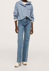 Persona che indossa un pullover azzurro chiaro con colletto, jeans blu a vita alta e gambe dritte, e sneakers bianche su uno sfondo semplice.