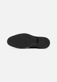 Semelle de chaussure noire avec grip texturé et talon bas, comportant l'empreinte du nom de la marque au centre.