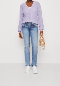 Ljuslila stickad cardigan med tre knappar, kombinerad med blå jeans och vita sneakers. Håller en liten genomskinlig väska med en tan-färgad rem.