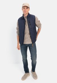 Marineblå puffervest med polstret tekstur, lynlåslommer, over en lysebrune sweater. Mørke jeans og beige sneakers fuldender outfittet.