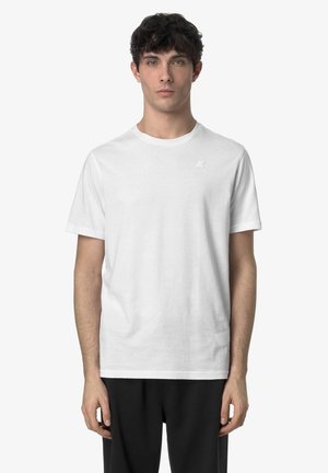 Giovane in piedi rivolto verso l'obiettivo, indossa una maglietta bianca a maniche corte semplice e pantaloni neri su uno sfondo bianco.