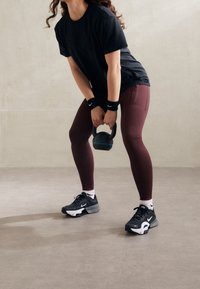 Černé tričko, vínové legíny, černobílé tenisky a náramky. Drží kettlebell, umístěný ve cvičební pozici na neutrální podlaze.