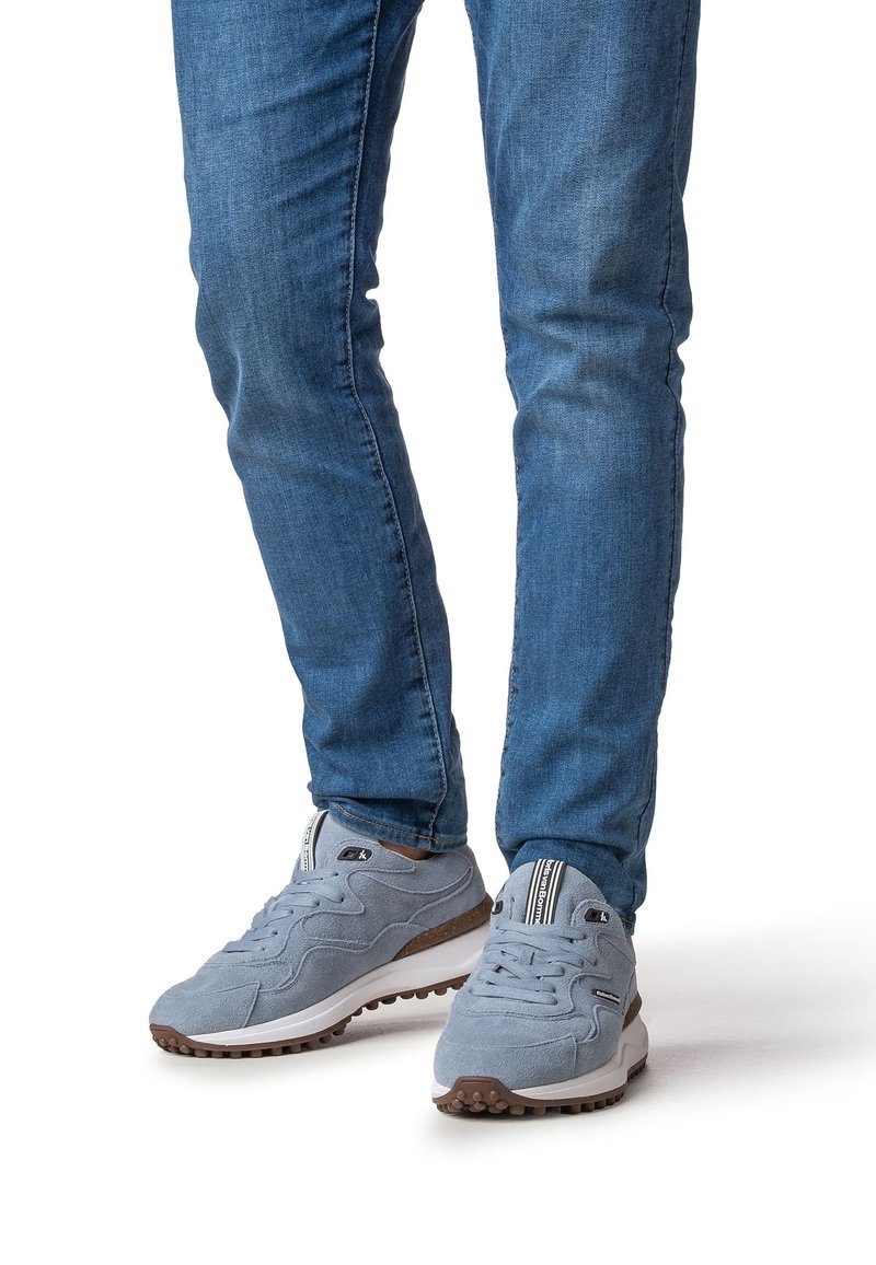 Grå ruskind sportssko med tekstureret overflade, brun gummiydersål og stribede stofdetaljer ved hælen, båret med blå jeans.