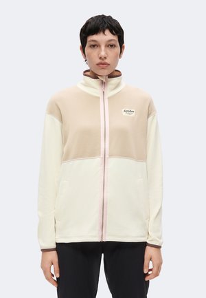 Jeune personne aux cheveux courts et foncés portant une veste polaire beige et crème zippée et un pantalon noir, debout devant un fond uni.