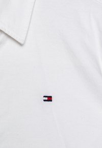 Hvit bomullspolo med en liten, brodert logo i marineblått, rødt og hvitt på venstre bryst. Klassisk kragedesign.