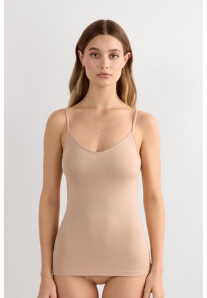 Joven mujer con una camiseta ajustada beige y ropa interior a juego, de pie frente a un fondo blanco liso, mirando hacia adelante.