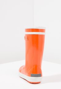 Pomarańczowy wodoodporny gumowy but z błyszczącym wykończeniem, białymi akcentami wokół góry i kontrastującą szaro-czarną pętelką na pięcie.